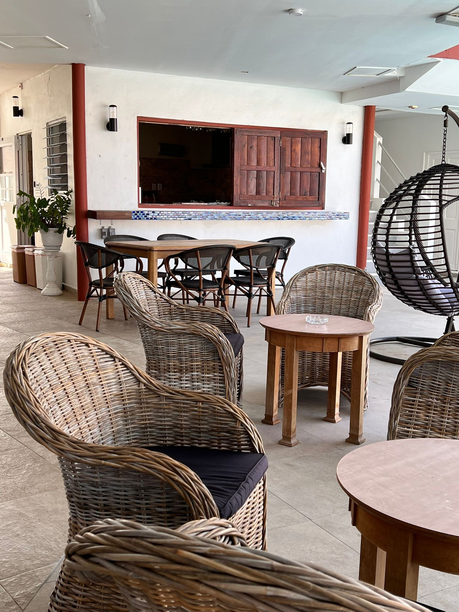 Un vaste espace extérieur aménagé avec des meubles en osier et une table en bois, agrémenté d’un mur recouvert de carreaux et de volets en bois.