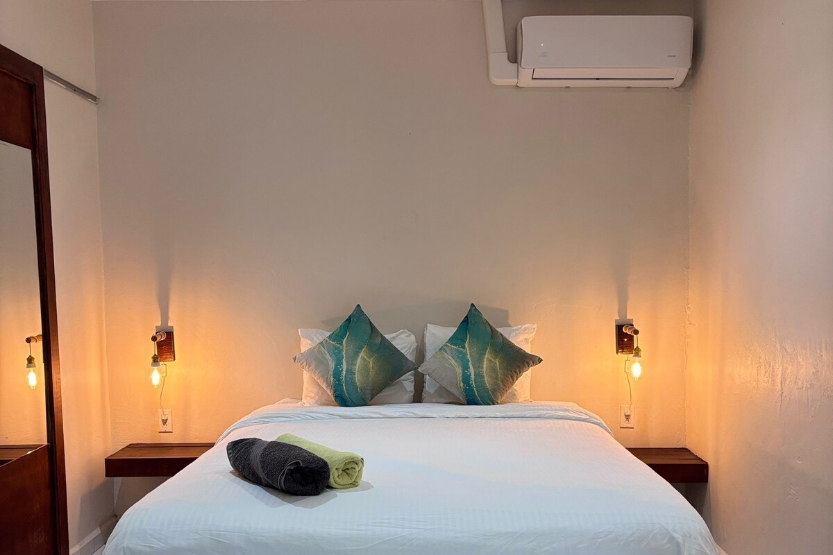 A hotel room with a bed, two nightstands, a wall-mounted air conditioner, and two wall sconces.