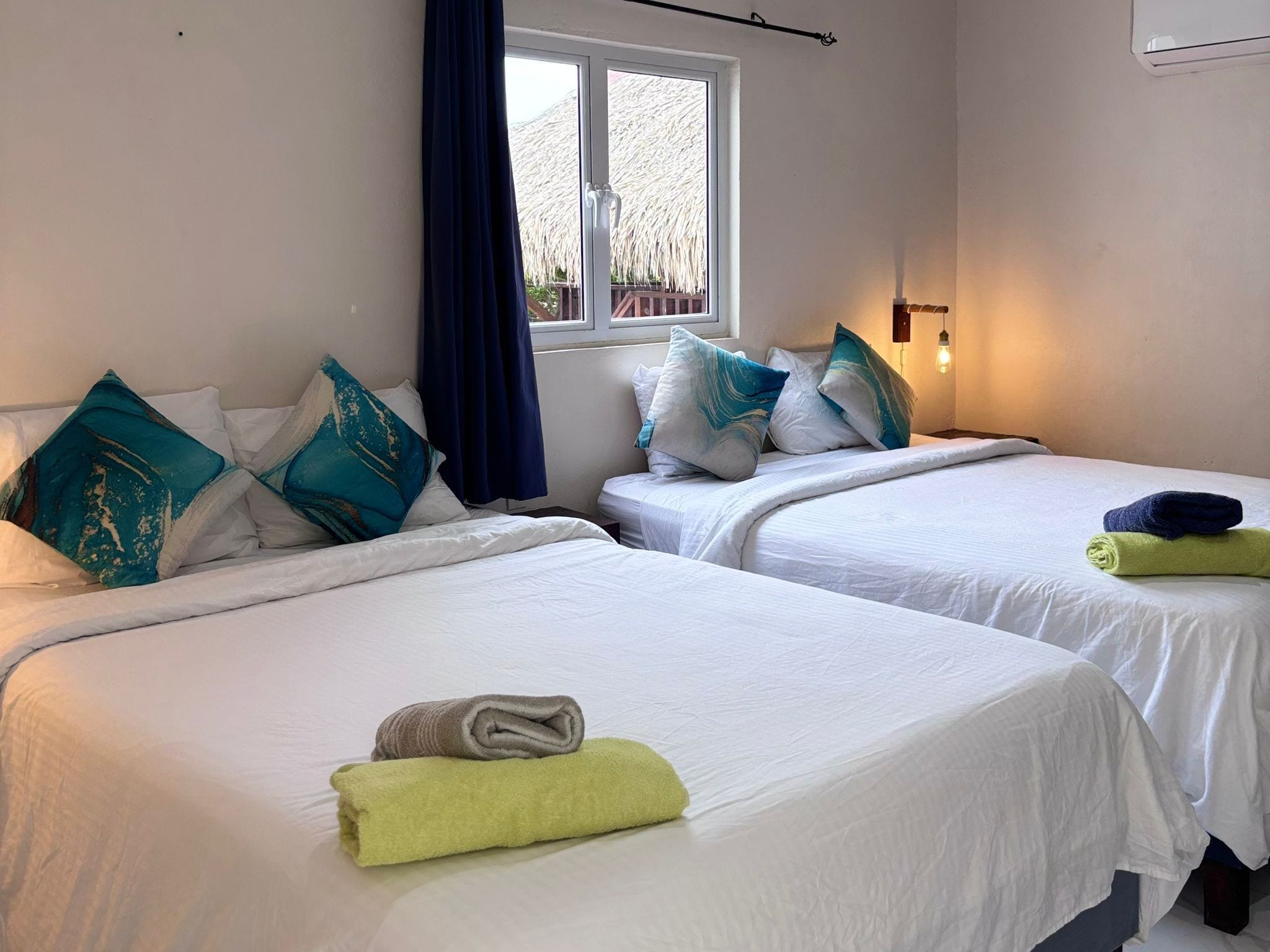 Een hotelkamer met twee bedden, elk voorzien van witte lakens en teal kussens. Er is een raam met uitzicht op een rieten dakstructuur.
