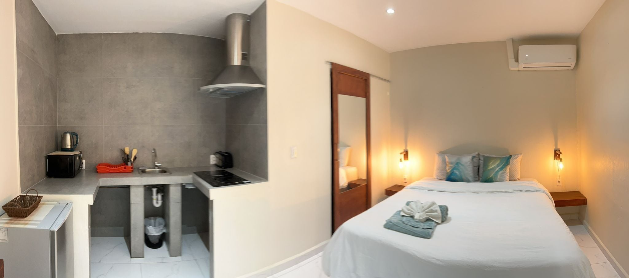 Een compacte hotelkamer met kitchenette, een bed met een netjes opgemaakt handdoekje en een nachtkastje met lamp.