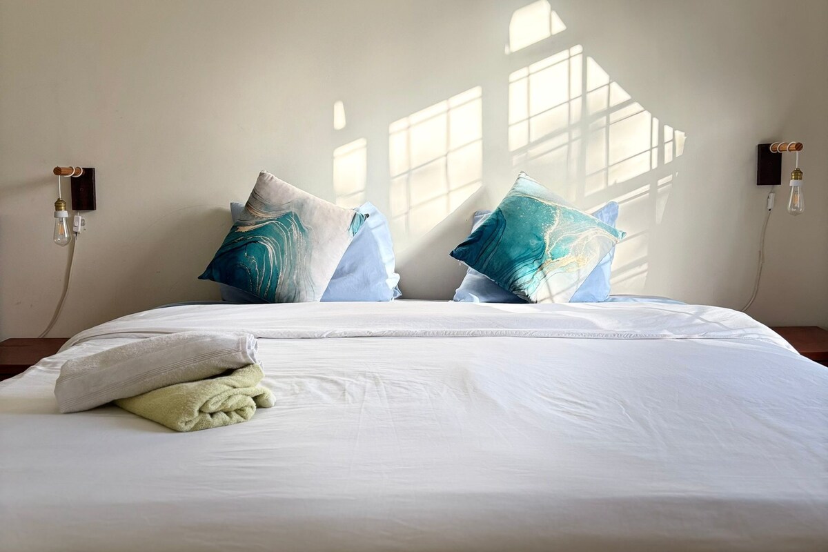 A neatly made bed with white linens, two decorative pillows with a blue and white pattern, and a folded green towel.