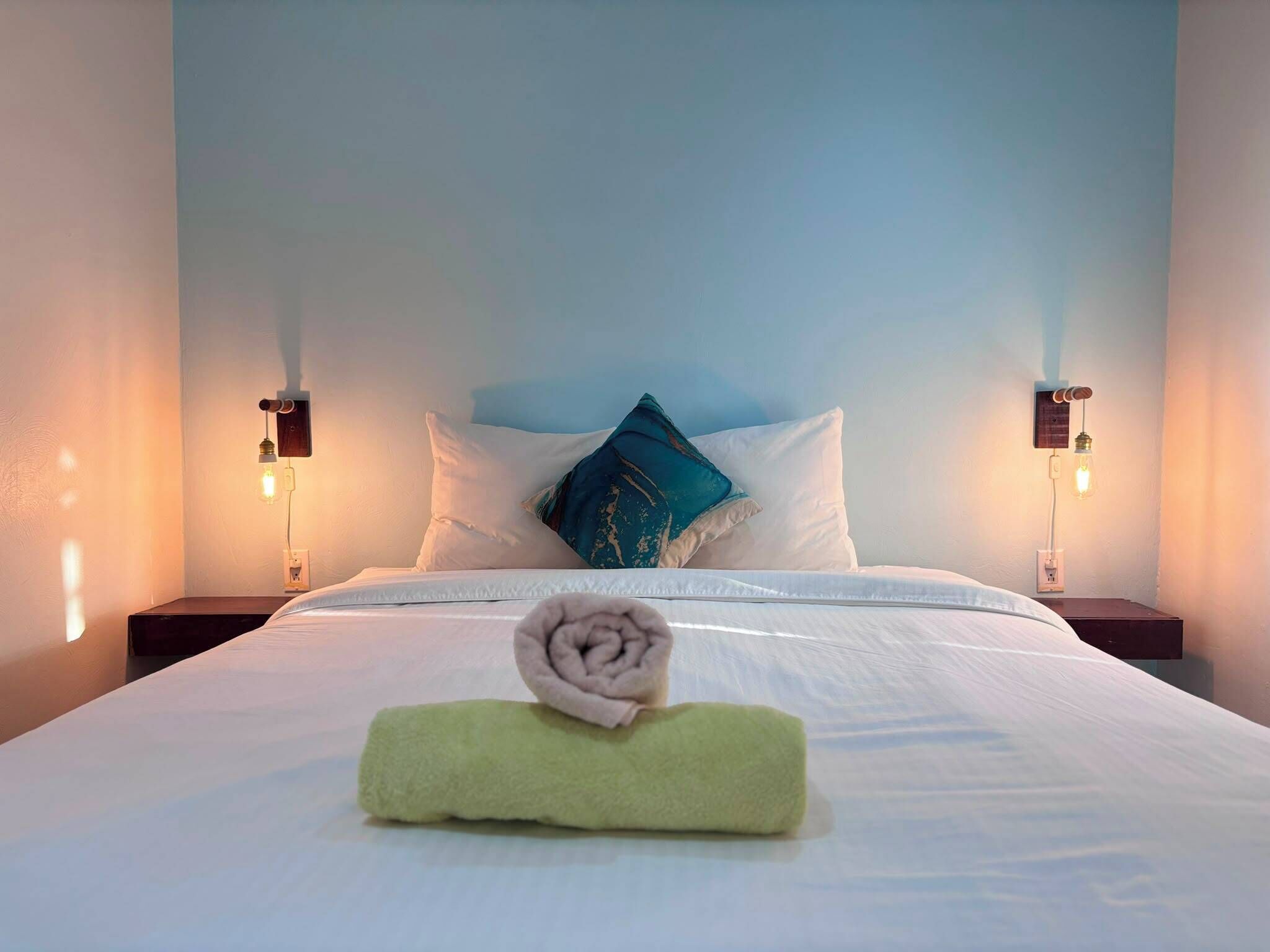A neatly made bed with white linens, a rolled towel, and a teal pillow. There are two bedside lamps on either side of the bed.
