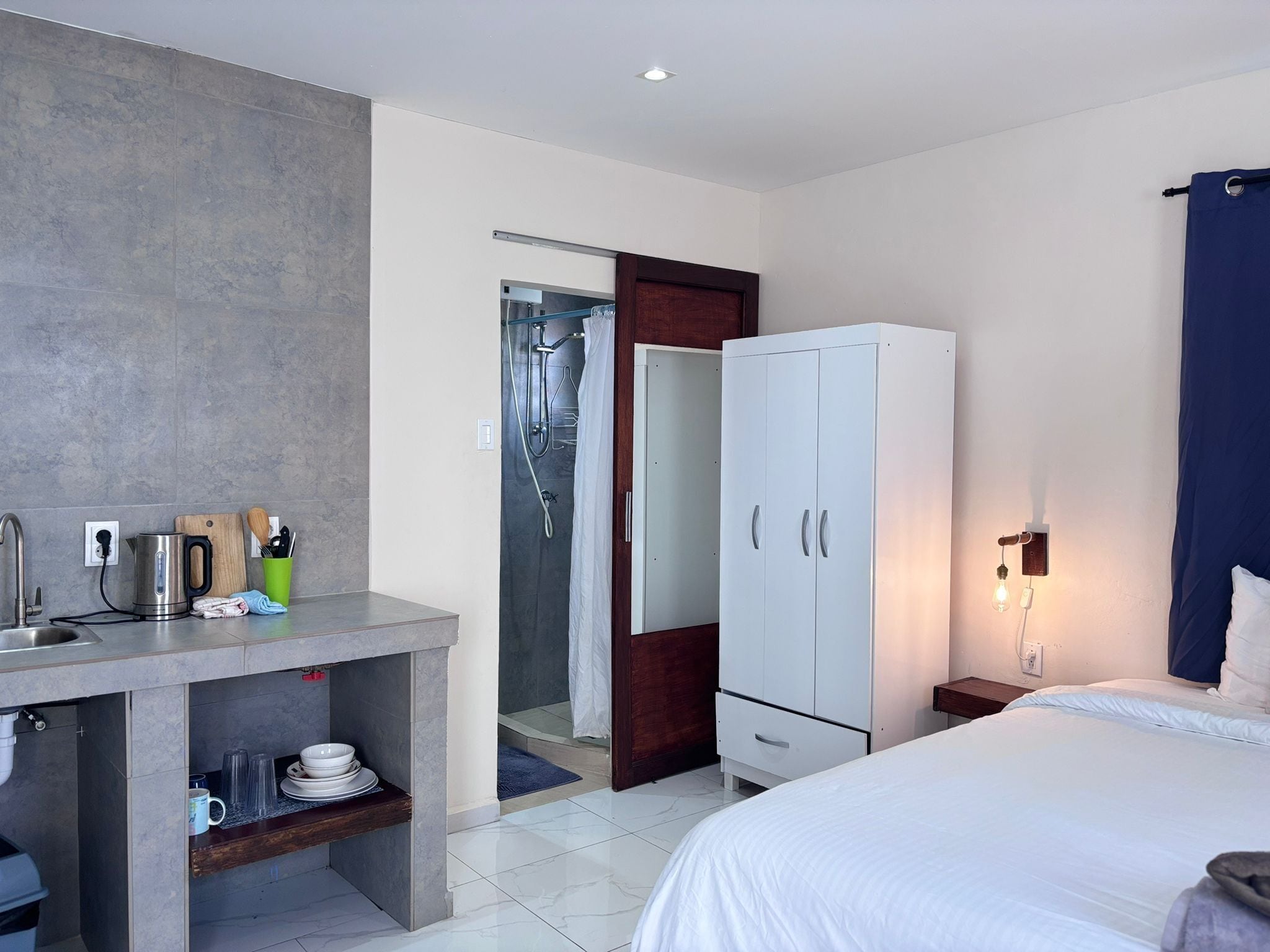 A modern hotel room with a bed, a desk with a kettle, a wardrobe, and a shower area.