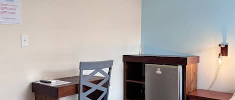 Quarto compacto com escrivaninha de madeira, cadeira, pequena geladeira, lixeira e luminária fixada na parede.
