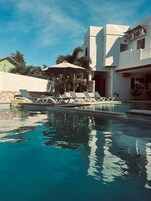 Piscina con sillones de descanso y una sombrilla, rodeada de edificios modernos y vegetación.