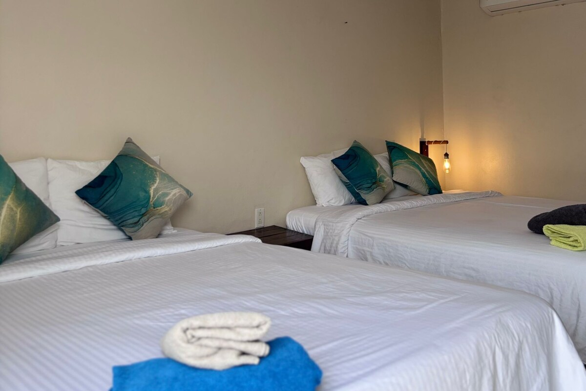 A hotel room with two beds, each with white bedding and teal pillows. There is a folded towel on the bed in the foreground.