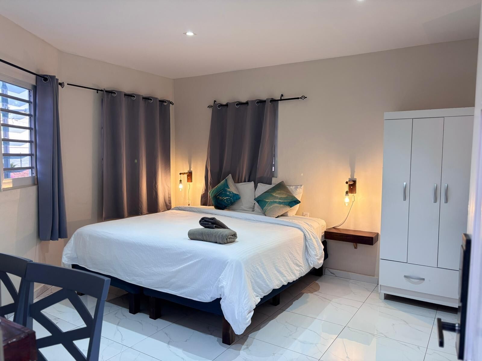 Un dormitorio con cama, armario, mesita de noche, silla y ventana con cortinas.