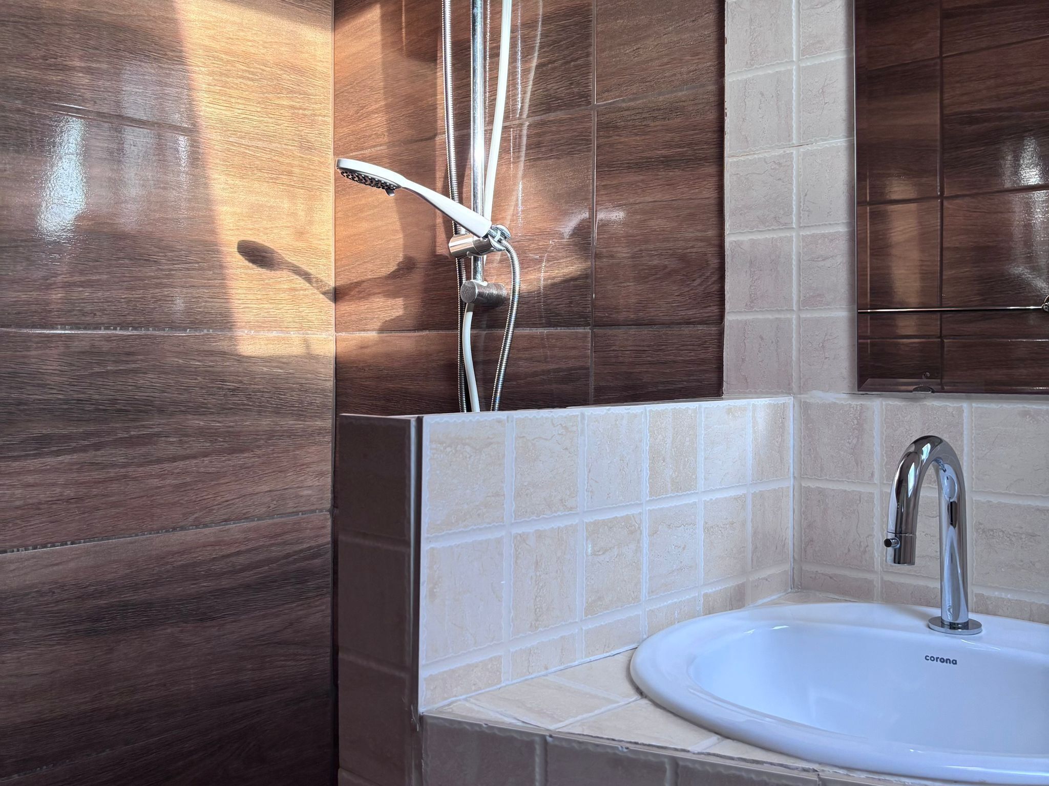 A bathroom with a white sink, chrome faucet, and a shower head mounted on a wall with brown tiles.