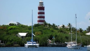 A lighthouse with a red and white striped tower, surrounded by lush greenery and a marina with sailboats.