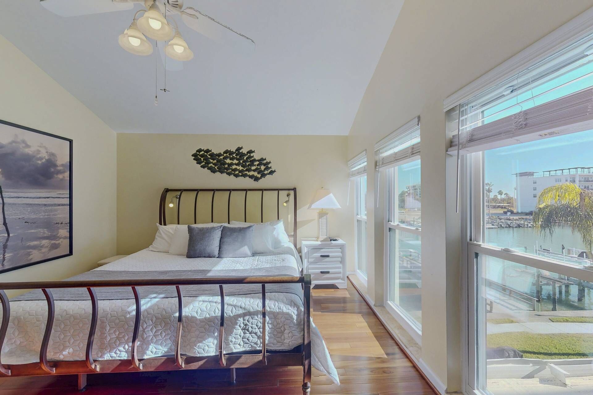A modern bedroom with a large bed, a ceiling light, a wall-mounted artwork, and a view of a marina through large windows.
