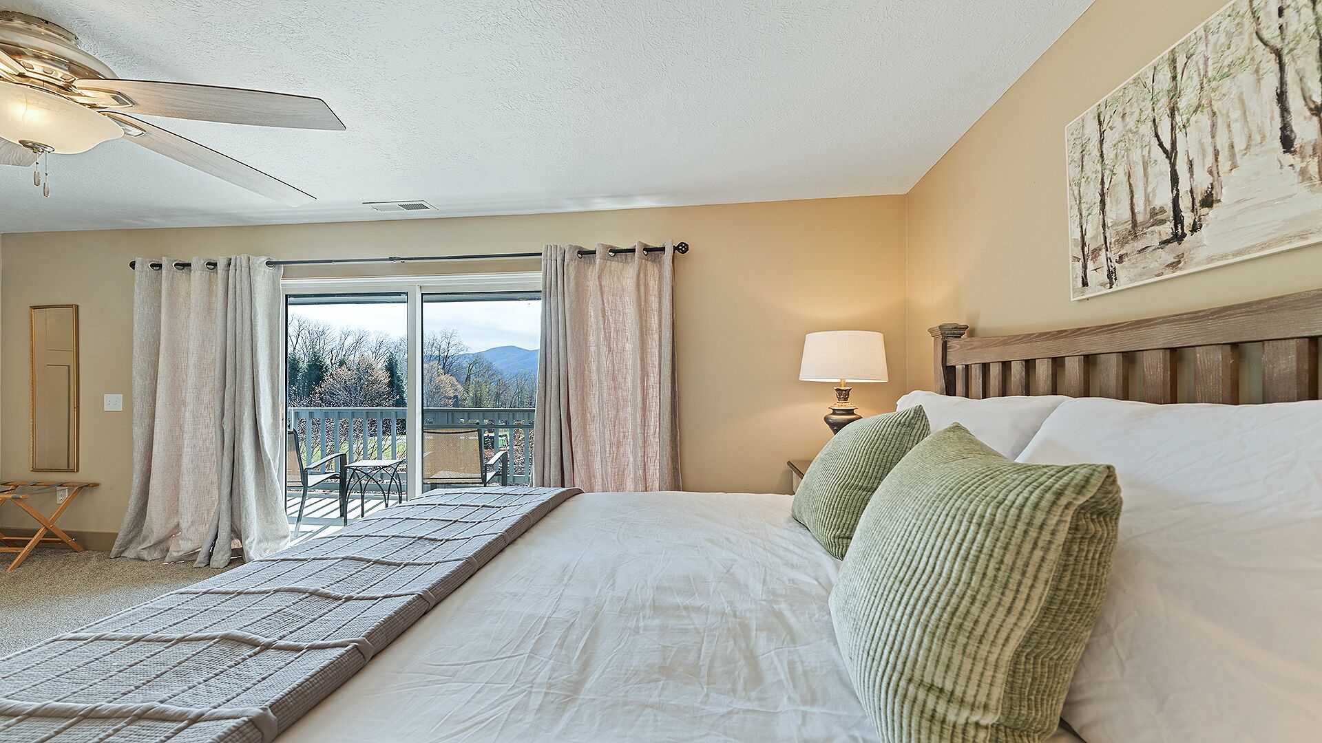 A bedroom with a bed, a nightstand, a lamp, a ceiling fan, a view through a glass door, and a painting on the wall.
