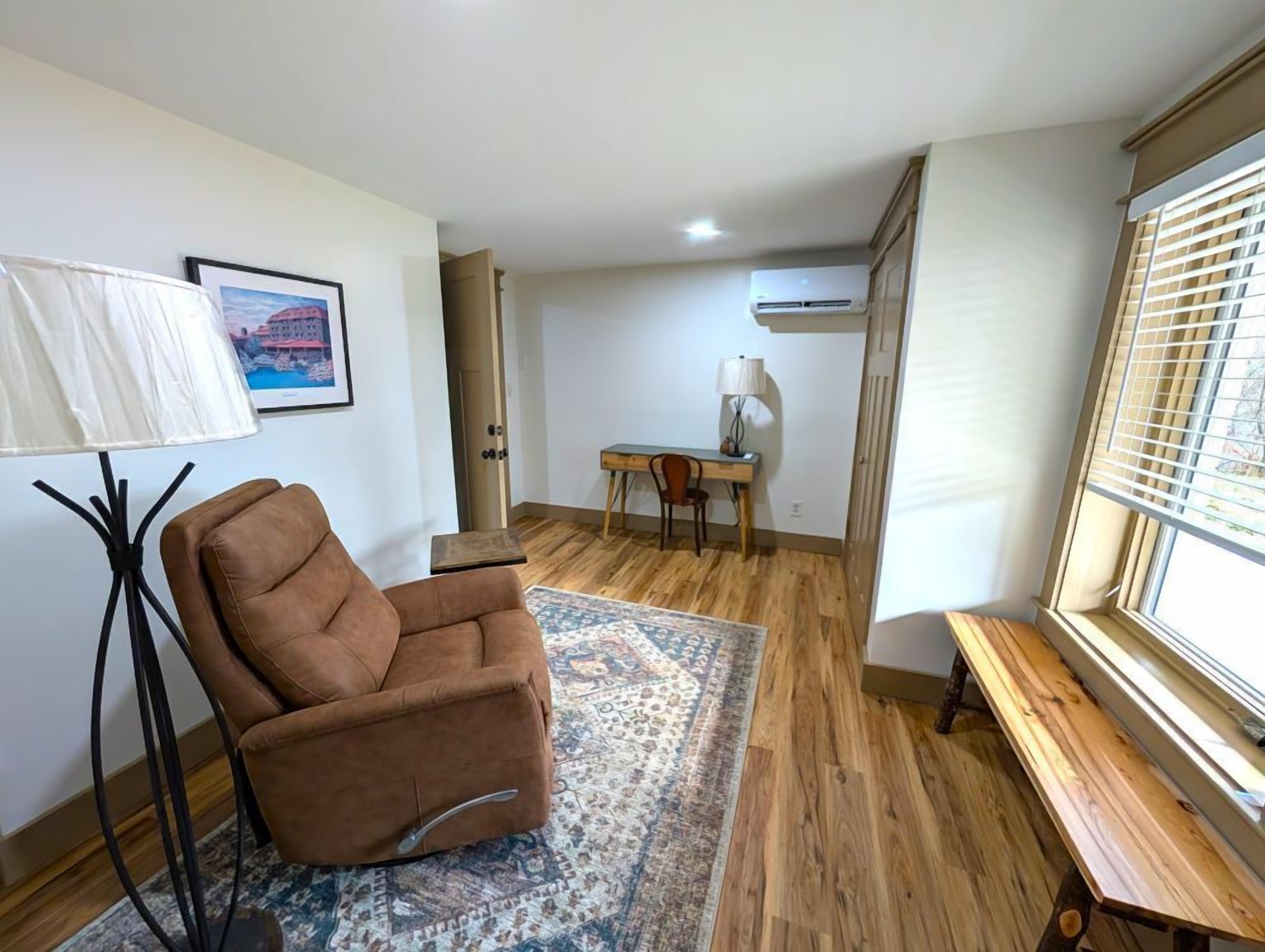 A room with a brown armchair, a small table, a lamp, a desk with a chair, a patterned rug, wooden flooring, an air conditioning unit, and a window with blinds.
