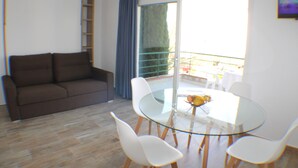 Un salon moderne avec un canapé marron, une table à manger en verre et des chaises blanches, ainsi qu’un balcon doté d’une balustrade.