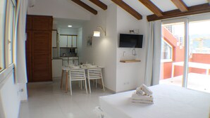 Une chambre d’hôtel avec un lit, un téléviseur fixé au mur, un coin repas avec des chaises et une table, et une cuisine à l’arrière-plan.