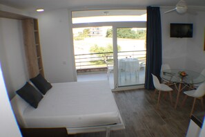 Ein Zimmer mit Bett, Tisch mit Stühlen, einem Balkon mit Tisch und Stühlen sowie einem Fernseher an der Wand.