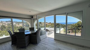 A balcony with a dining table and chairs, large arched windows, and a view of a coastal town. - Villa Luna del mar Su (Benissa)
