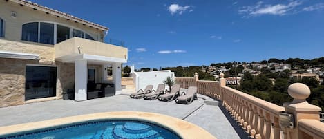 A pool area with a clear blue pool, lounge chairs, and a view of a hillside town.
