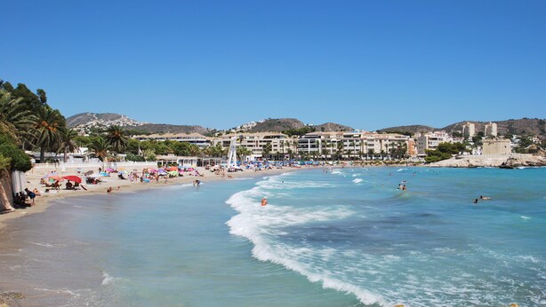 A beach with clear blue water, palm trees, and a resort area with buildings in the background. - Villa Luna del mar Su (Benissa)