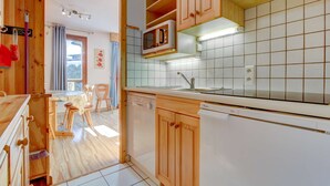A kitchen with wooden cabinets, a microwave, a dishwasher, and a dining area with chairs and a table. - Fanyon 9 : Central one bedroom + sleeping area (MORZINE)