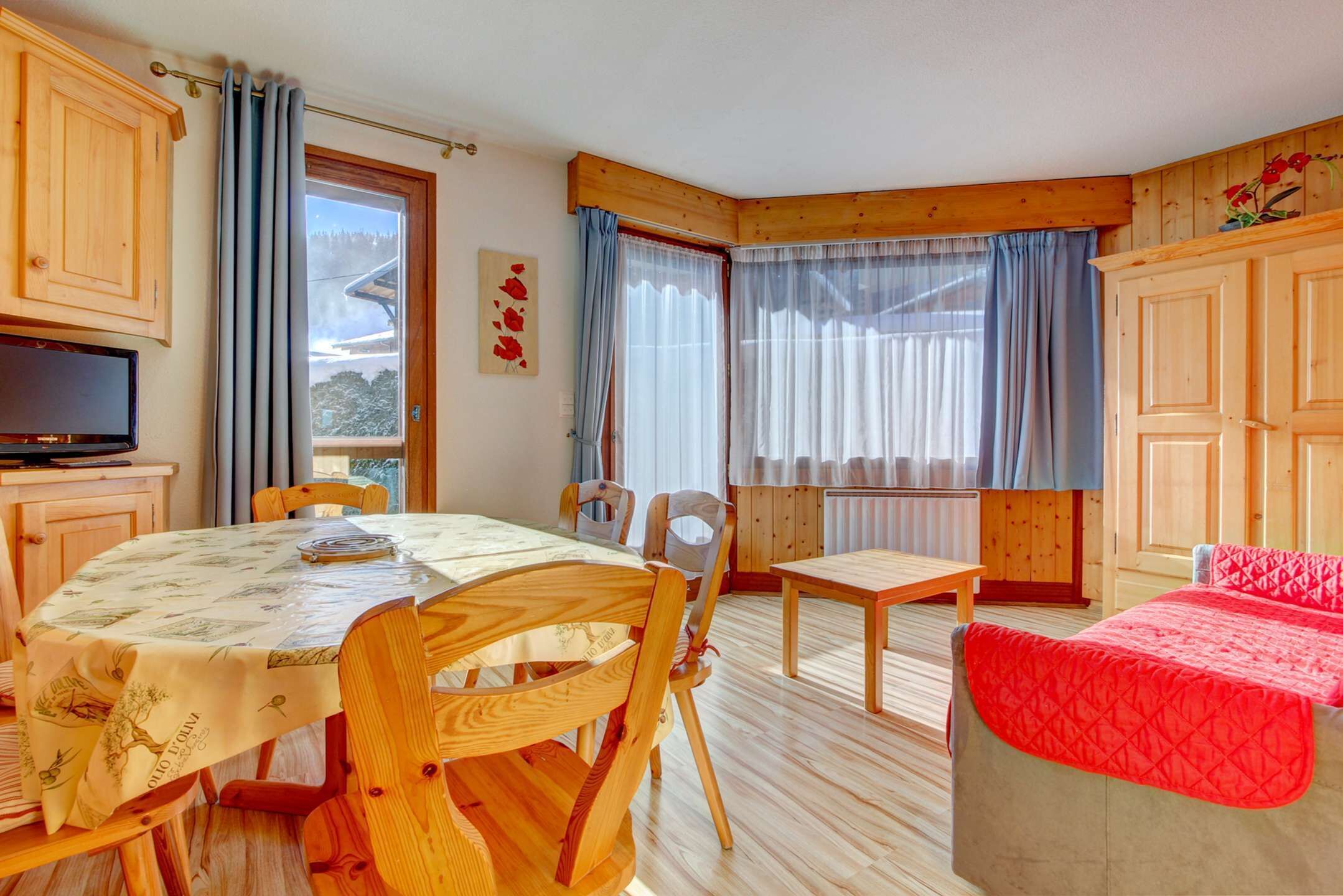 A room with a wooden dining table, chairs, a bed with a red quilt, a television, and a window with curtains.