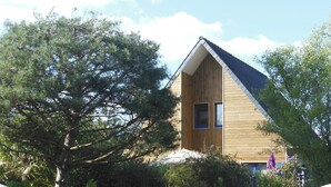 A wooden house with a sloped roof, surrounded by dense greenery and a gravel driveway. - Pretty Cozy 3 * house, green 7/8 pers St Gildas de Rhuys, 1km bg, 2km beach (Saint-Gildas-De-Rhuys)