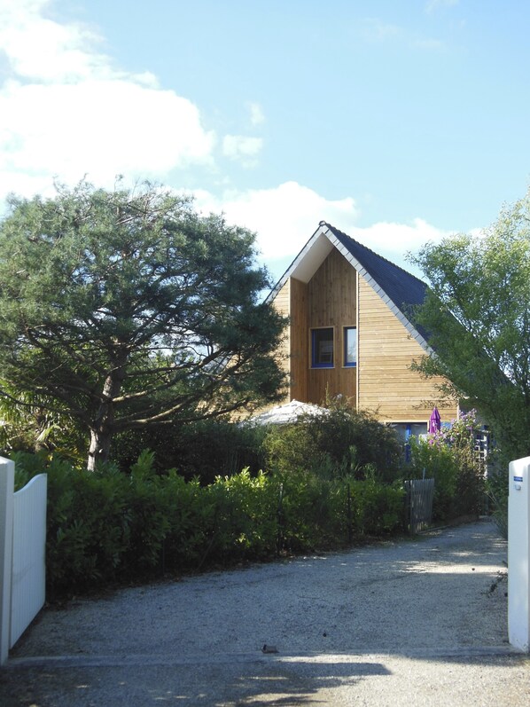 A wooden house with a sloped roof, surrounded by dense greenery and a gravel driveway.