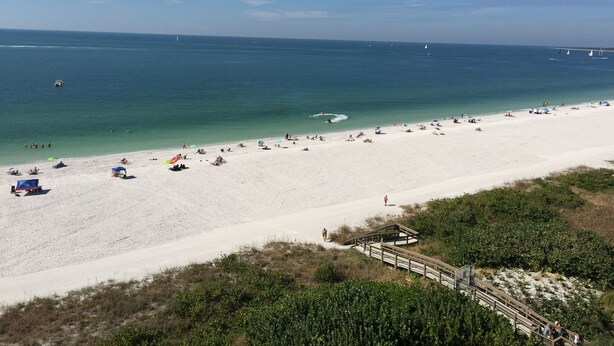 A beach with a wooden boardwalk leading to the shore, clear blue water, and a few sailboats in the distance.