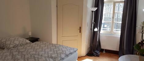 A bedroom with a bed, a nightstand, a lamp, a window with curtains, and a view of buildings outside.