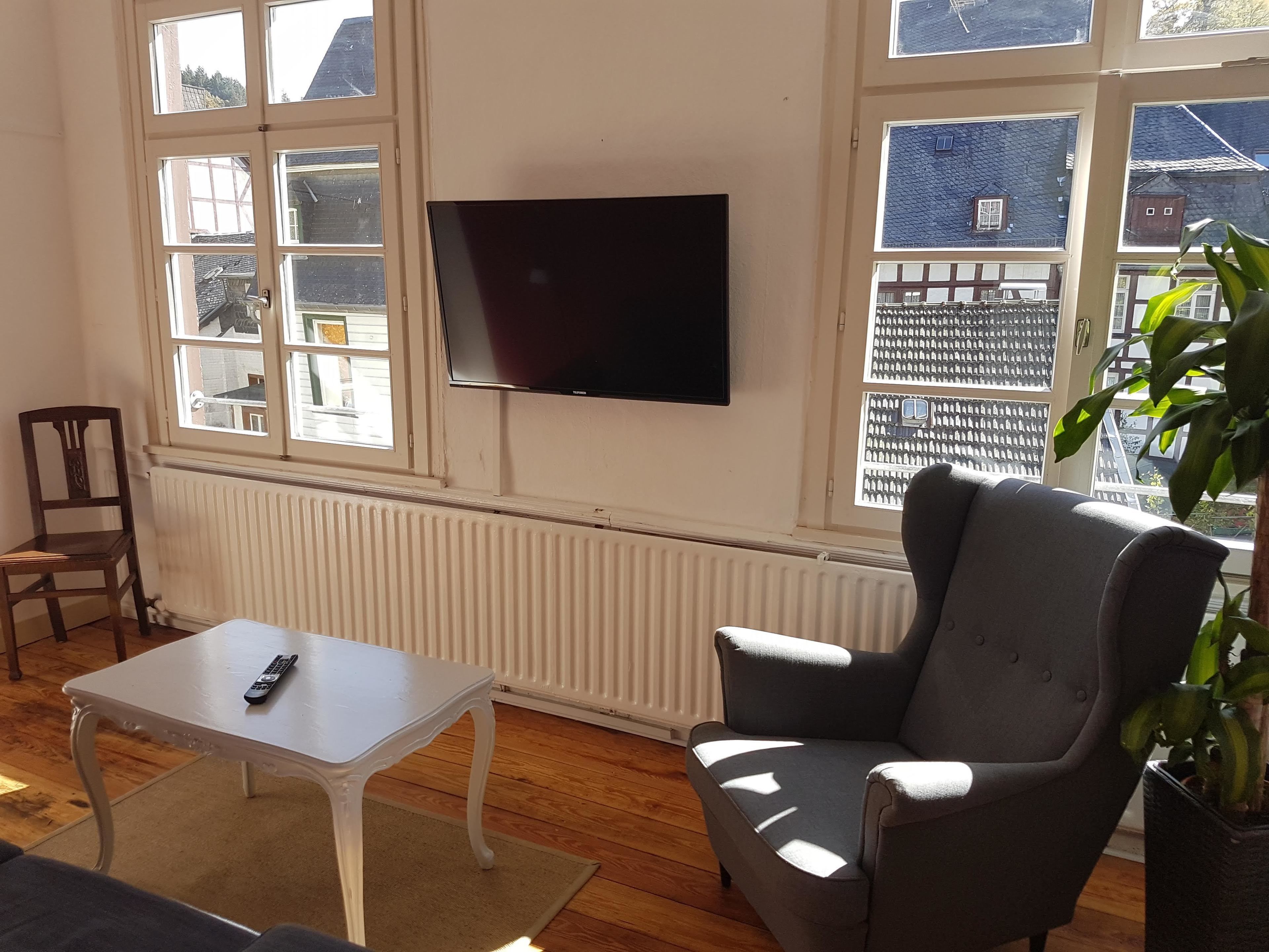 A room with a large window, a flat-screen TV mounted on the wall, a grey armchair, a white table, and a potted plant.