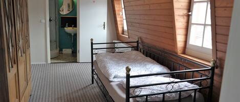 A small, single-bed room with a metal frame, a wooden wardrobe, a bathroom with a mirror and sink, and a sloped ceiling with wooden paneling.