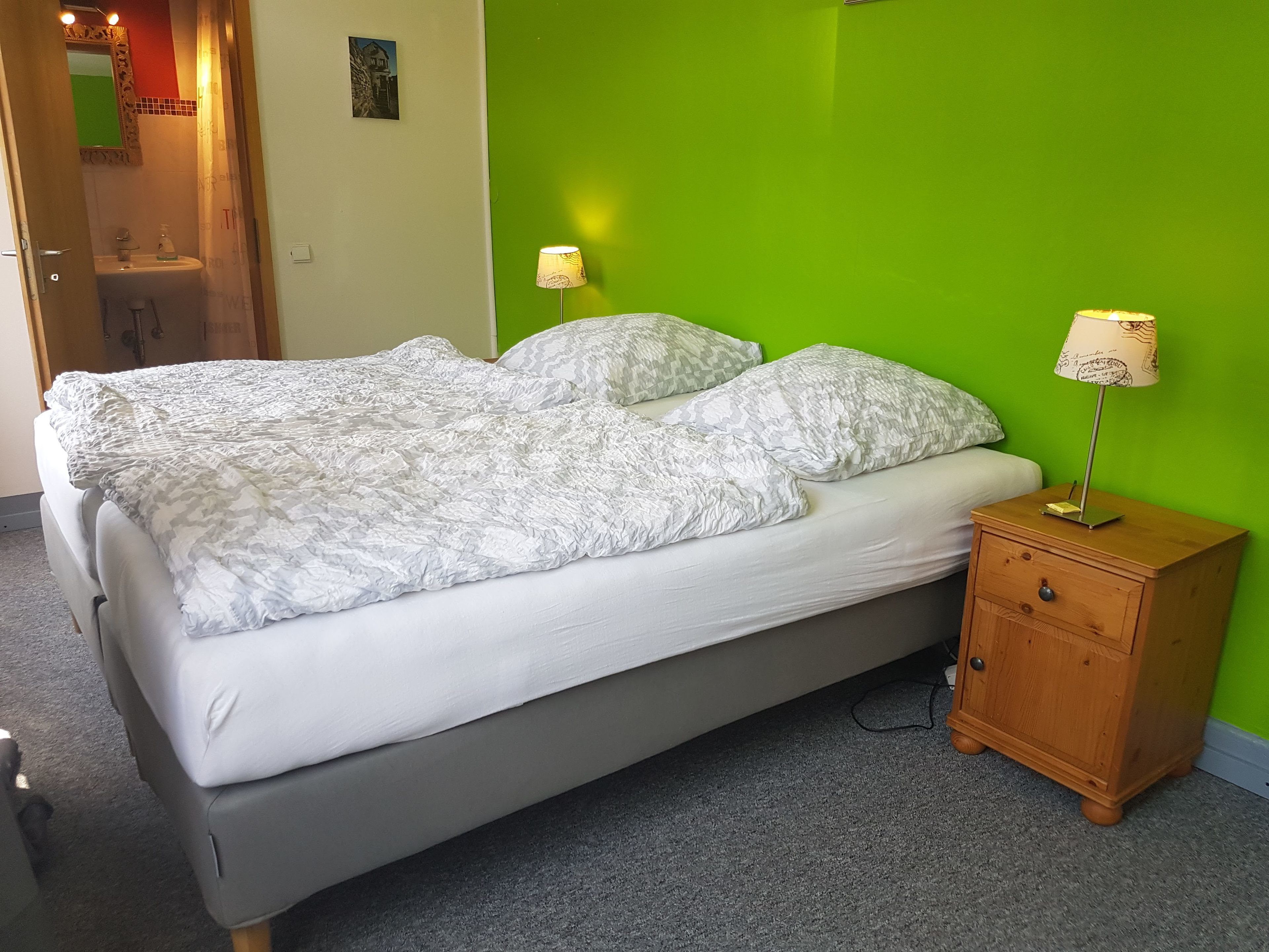 A bed with a white comforter and pillows, a wooden nightstand with a lamp, and a green wall.