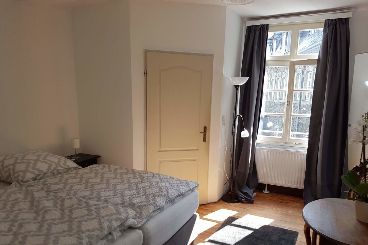A bedroom with a bed, a nightstand, a window with curtains, a radiator, and a small table with a plant.