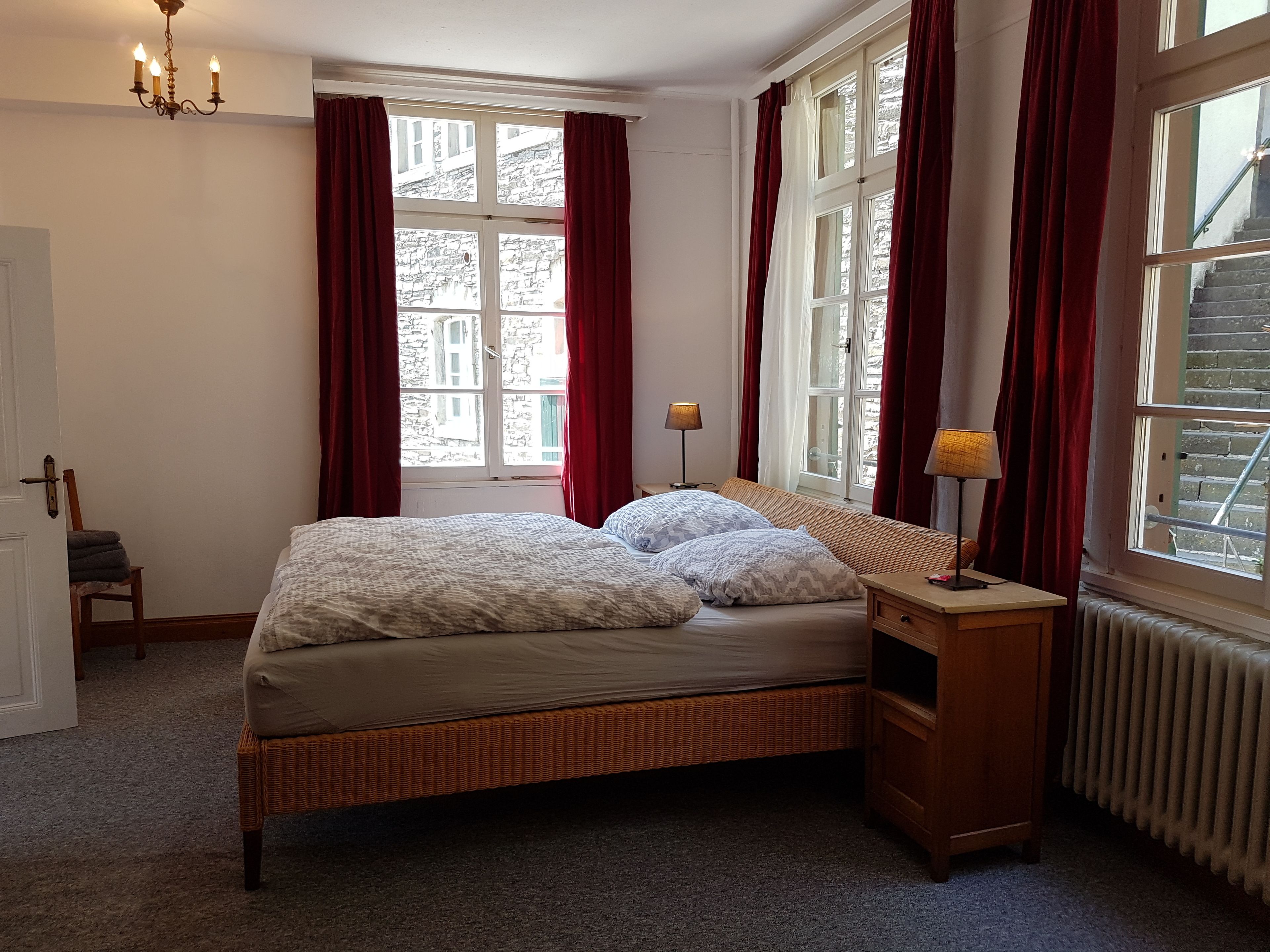Une chambre à coucher bien aménagée, avec un grand lit, une table de chevet en bois, une lampe et une vue sur la ville grâce à de grandes fenêtres ornées de rideaux rouges.