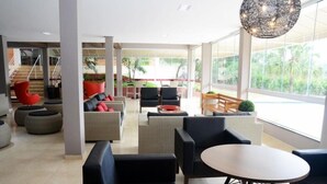 A modern lounge area with sofas, chairs, and a round table, large windows with a view of greenery, and a staircase in the background. - Hotel Giordano Mantiqueira (Sao Joao da Boa Vista)