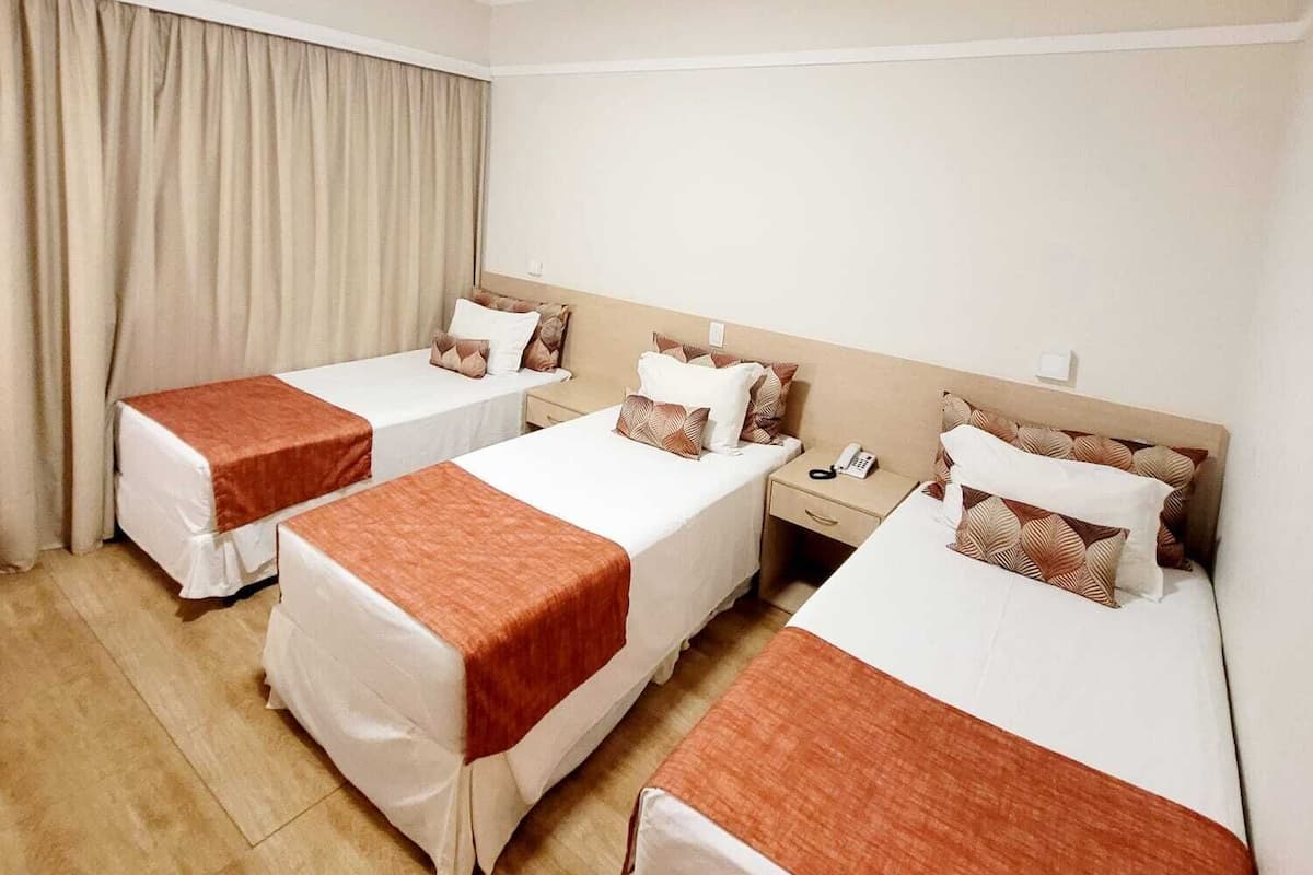 Une chambre d’hôtel avec trois lits simples, chacun recouvert d’une courtepointe blanche et marron, doté d’une tête de lit en bois et d’une table de chevet avec un téléphone.