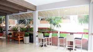A hotel lobby with a wooden bench, white chairs, and a glass wall offering a view of the outdoors. - Hotel Giordano Mantiqueira (Sao Joao da Boa Vista)