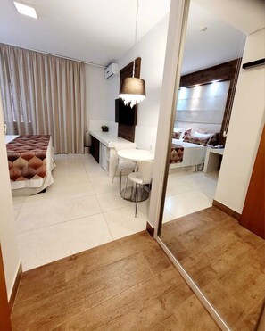 Una habitación de hotel moderna con cama, escritorio y zona de estar. Cuenta con un espejo grande en una pared y suelo de baldosas.