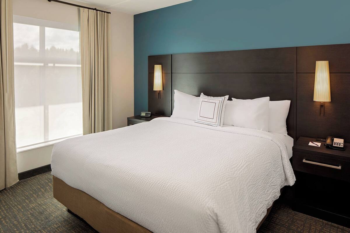 Una habitación de hotel con una cama grande, dos mesitas de noche, una lámpara y un ventanal con cortinas.
