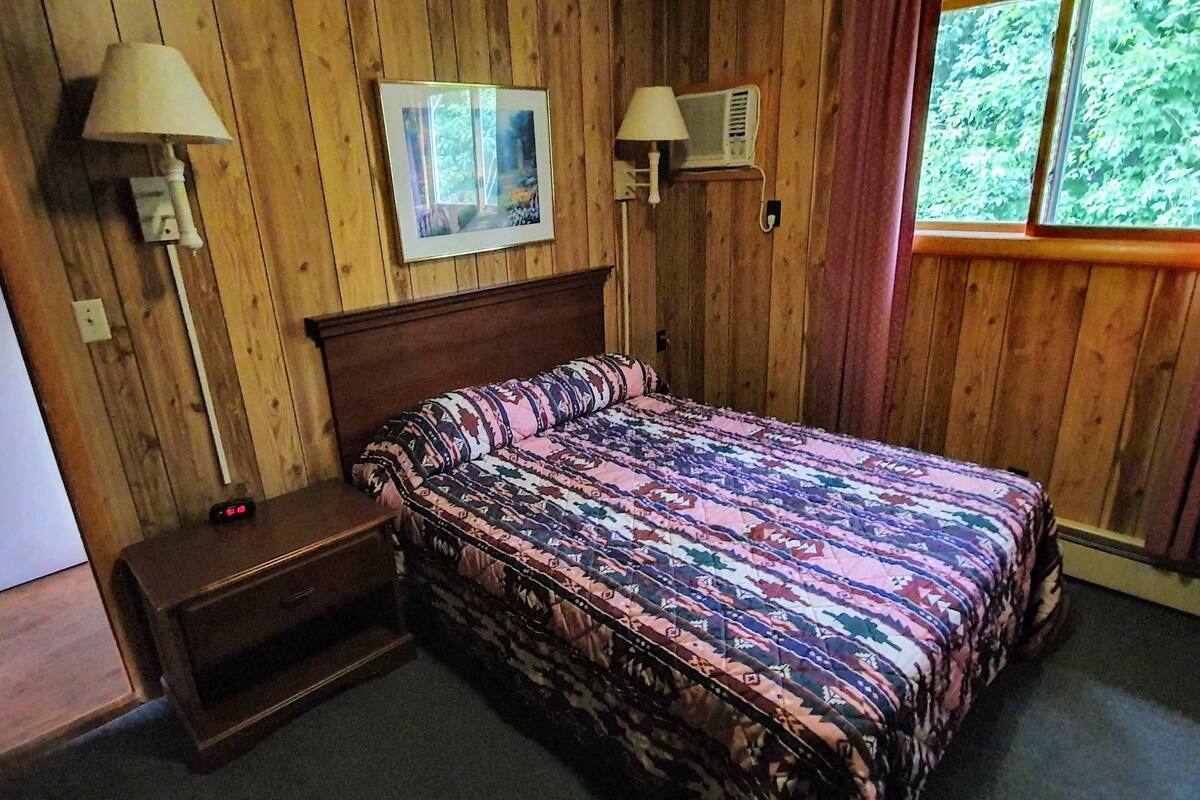 A wooden-paneled room with a bed, two wall-mounted lamps, a picture, and a window with curtains.