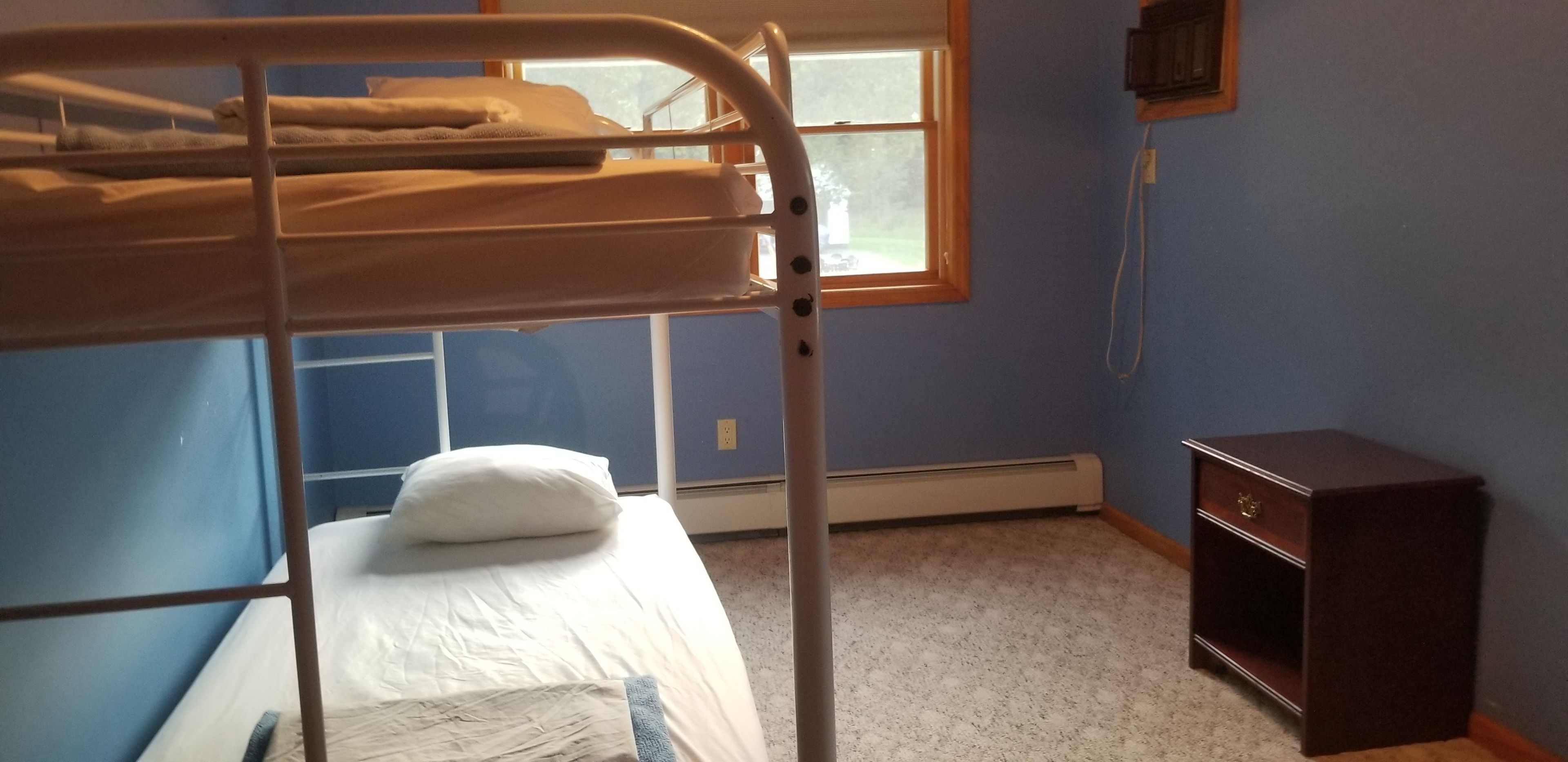 A bunk bed with a white mattress and a pillow, a small wooden nightstand, and a window with blinds.