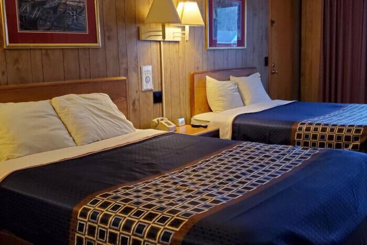 A hotel room with two beds, wooden paneling, a nightstand with a lamp, and framed artwork on the walls.