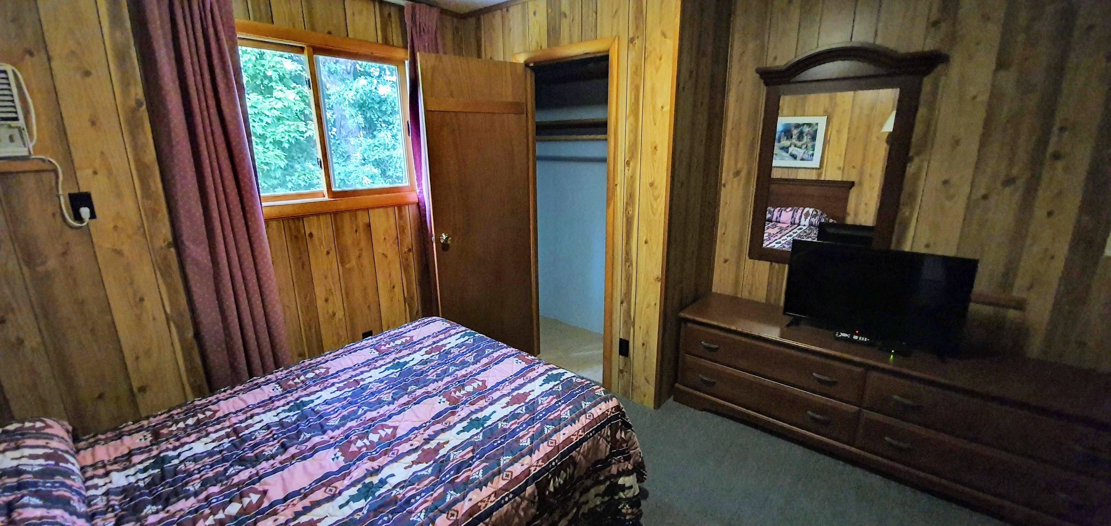 A wooden-paneled room with a bed, a dresser, a television, and a window with curtains.