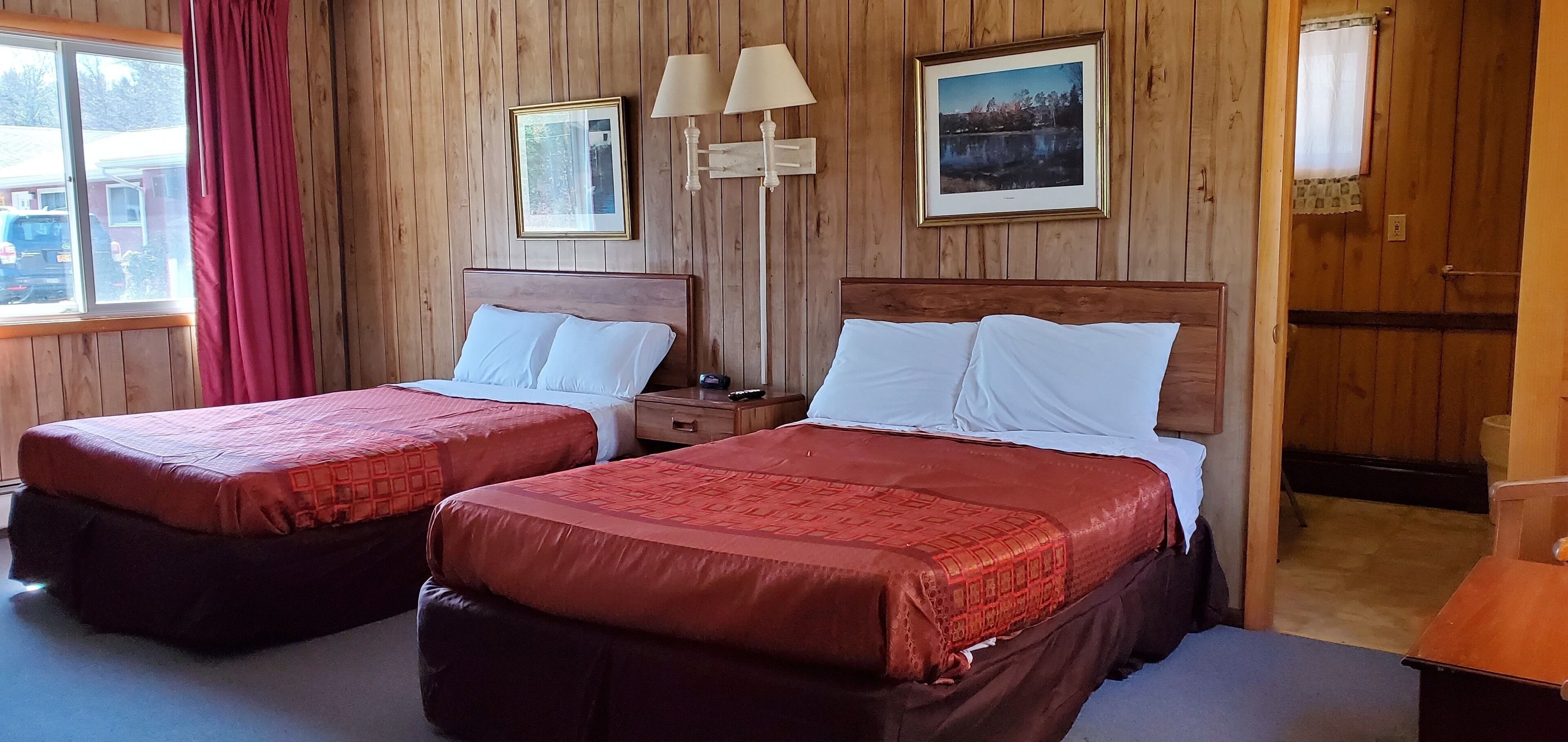 A room with two beds, wooden walls, a window with a view, and a nightstand with a lamp.