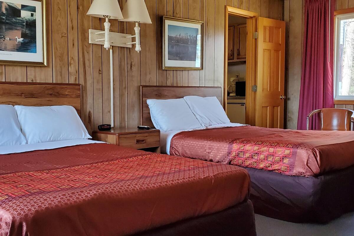 A room with two beds, wooden walls, a desk with a lamp, and a window with curtains.