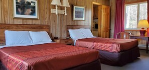 A room with two beds, wooden walls, a desk with a lamp, and a window with curtains.
