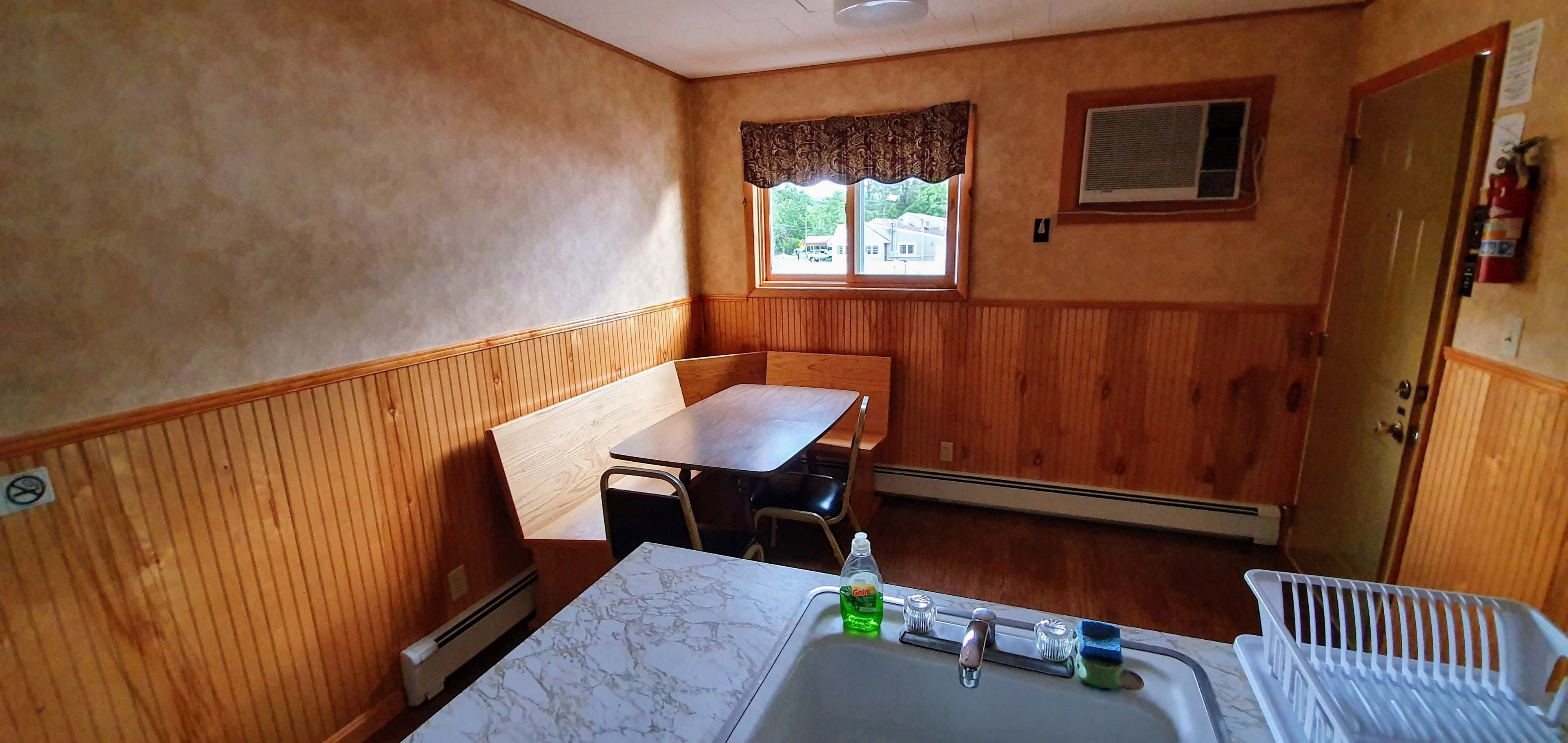 Une petite pièce accueillante aux murs lambrissés, avec une seule table et une seule chaise, un lavabo avec un miroir et une fenêtre agrémentée d’une lambrequin décorative.
