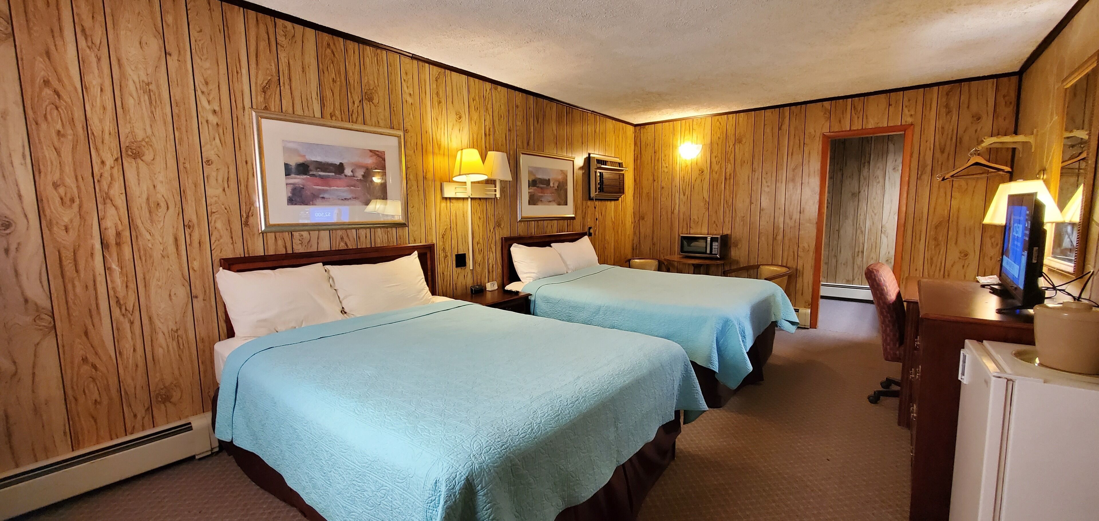 A hotel room with wooden panel walls, two beds with light blue bedding, a desk with a computer, a microwave, and a small table with a chair.