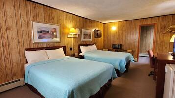 A hotel room with wooden panel walls, two beds with light blue bedding, a desk with a computer, a microwave, and a small table with a chair.
