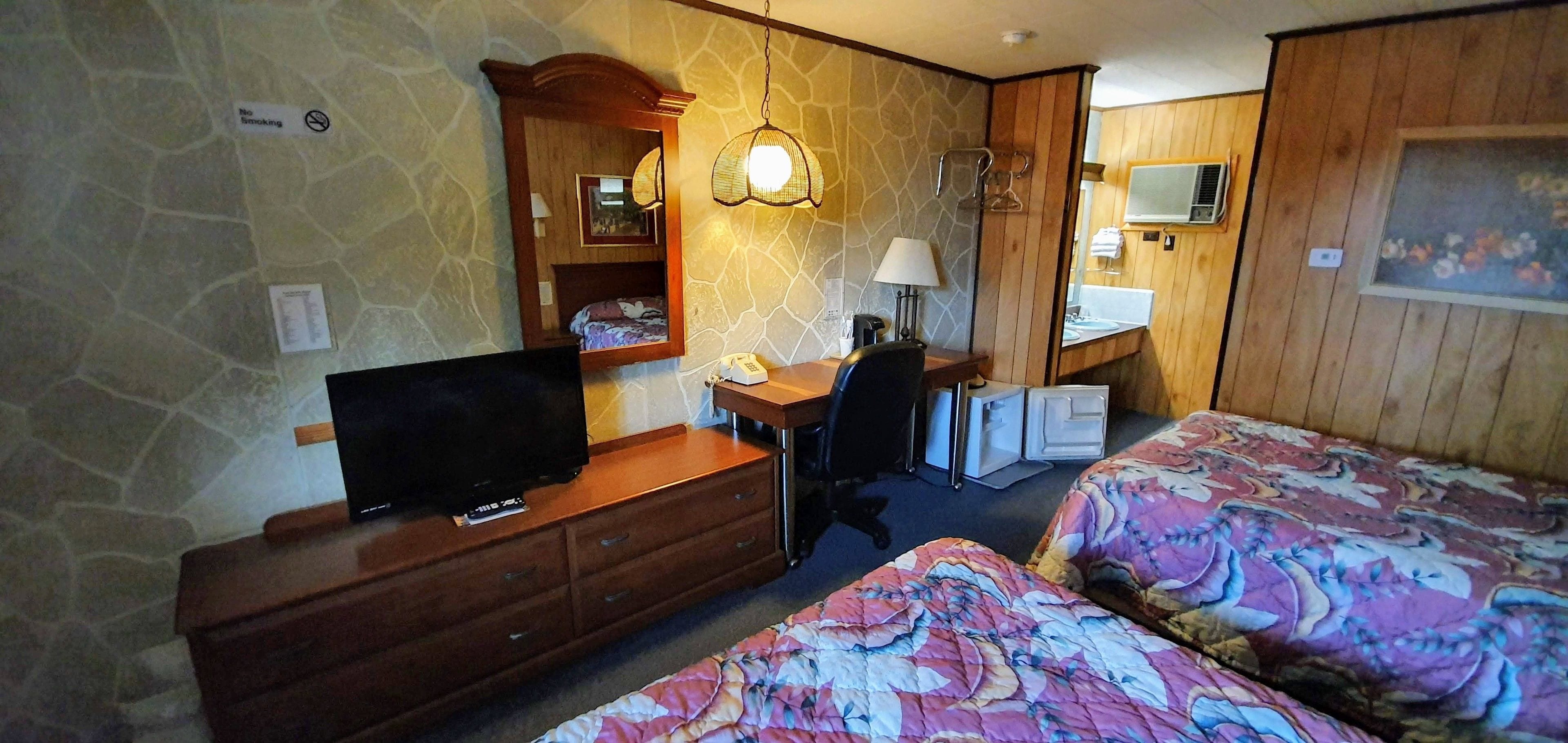 Une chambre d’hôtel avec un mur en pierre, un bureau en bois, une télévision, un lit avec une literie à motifs floraux, un miroir et une petite kitchenette.