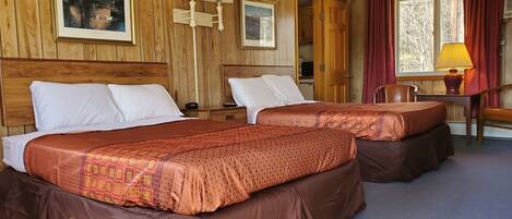A hotel room with two beds, wooden walls, a desk with a lamp, and a window with curtains.